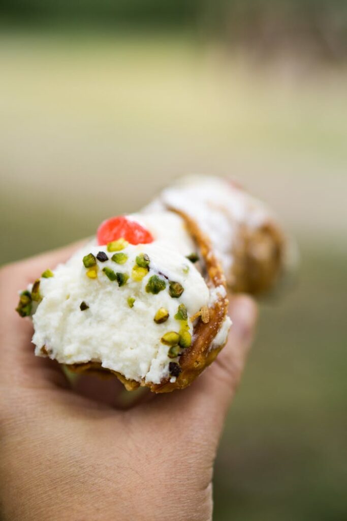 Close-up of a hand holding a creamy cannoli topped with pistachios and cherry, outdoors.